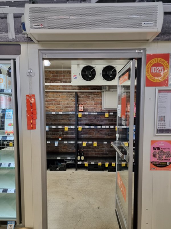 An air curtain mounted over a cool room door in a bottle shop, the door standing open onto shelving inside the cool room