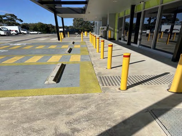 A line of yellow bollards along a glass shopfront, beside a tactile strip